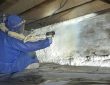Worker installing closed-cell spray foam insulation under house floor joists in crawl space