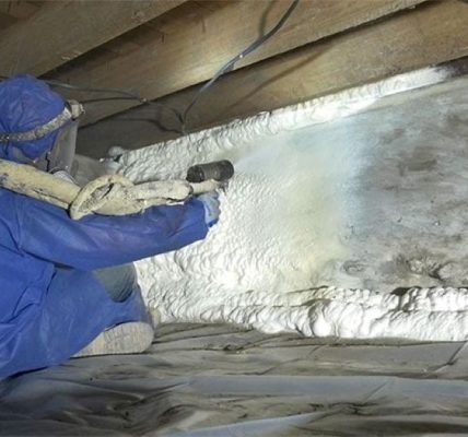 Worker installing closed-cell spray foam insulation under house floor joists in crawl space