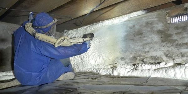 Worker installing closed-cell spray foam insulation under house floor joists in crawl space