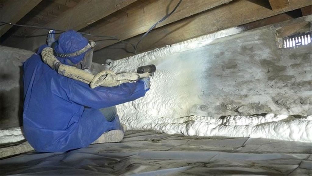 Worker installing closed-cell spray foam insulation under house floor joists in crawl space