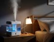 Modern white large capacity humidifier with illuminated water tank on nightstand beside bed in dark bedroom producing cool mist